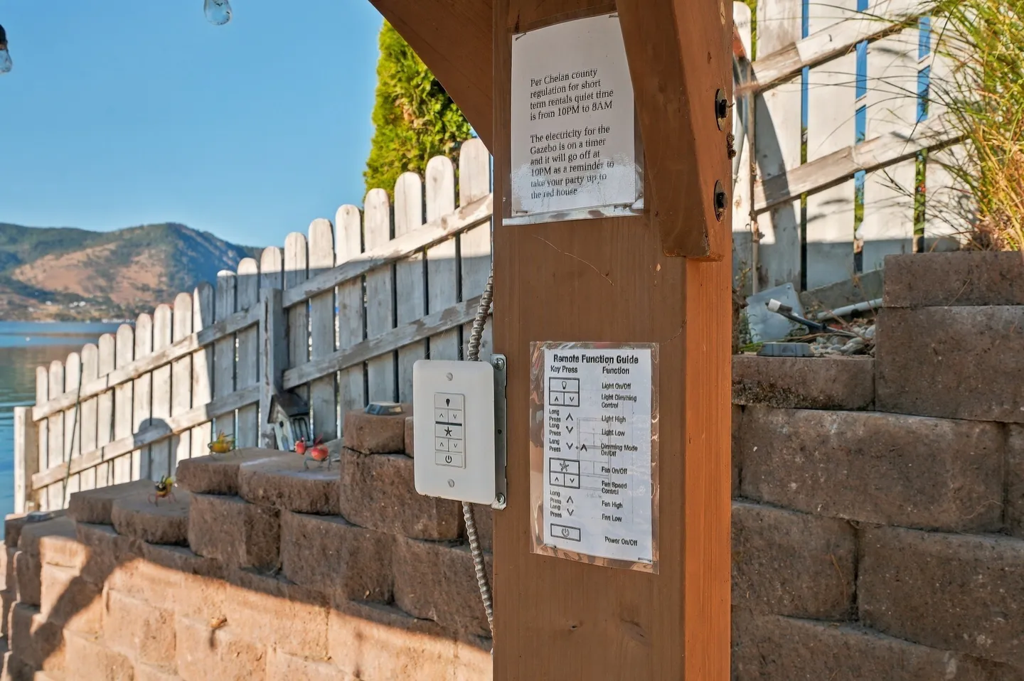 Close-up of a wooden gazebo post with posted house rules and remote control instructions, overlooking a lake and fence.