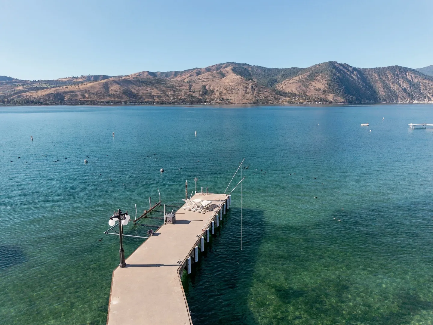 An elevated view of a concrete dock extending into clear turquoise lake water with mountains across the bay.