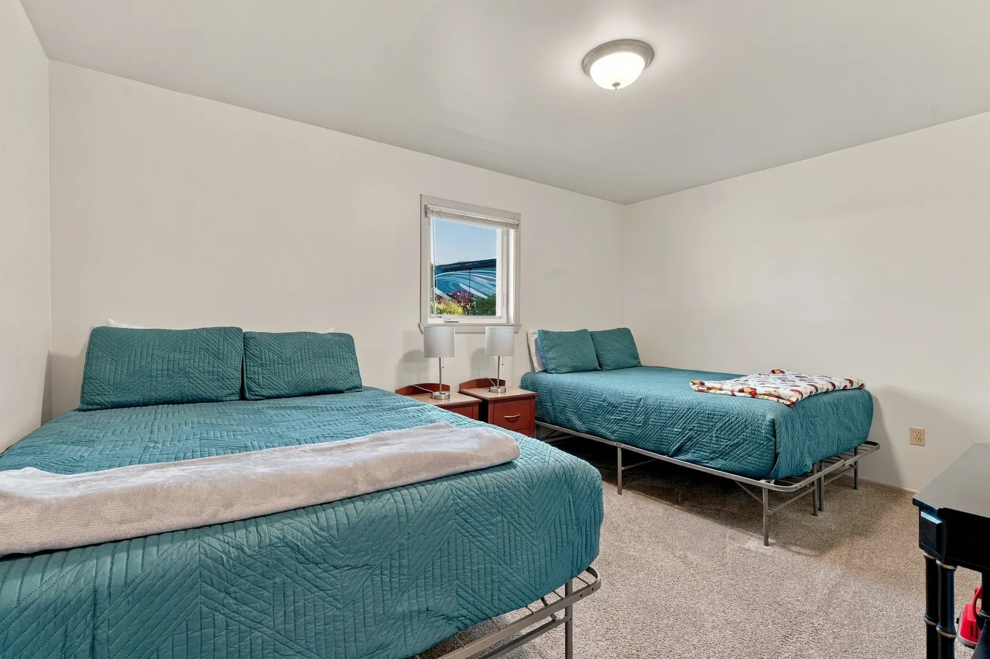 Bedroom featuring two beds with teal quilts, metal frames, carpeted flooring, and a small window between two nightstands.