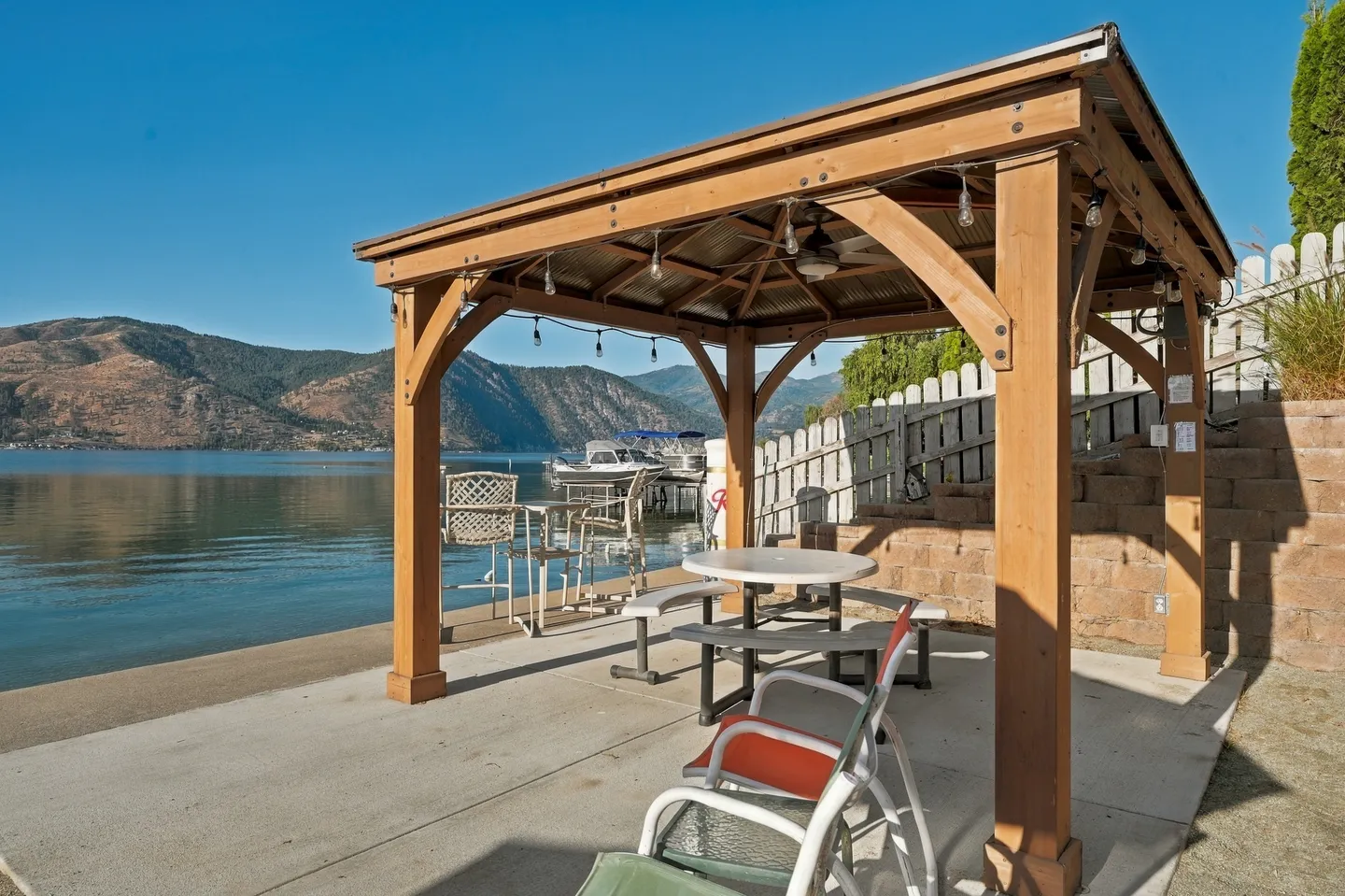 A wooden gazebo with outdoor seating sits on a concrete patio beside a lake with mountains in the background.