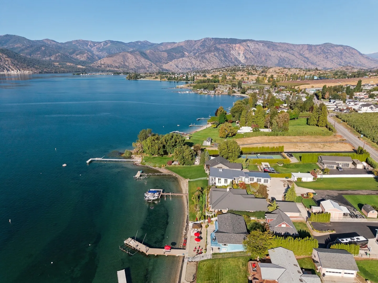 Aerial view of a lakefront community with private docks, houses, and mountains in the distance under a clear sky.