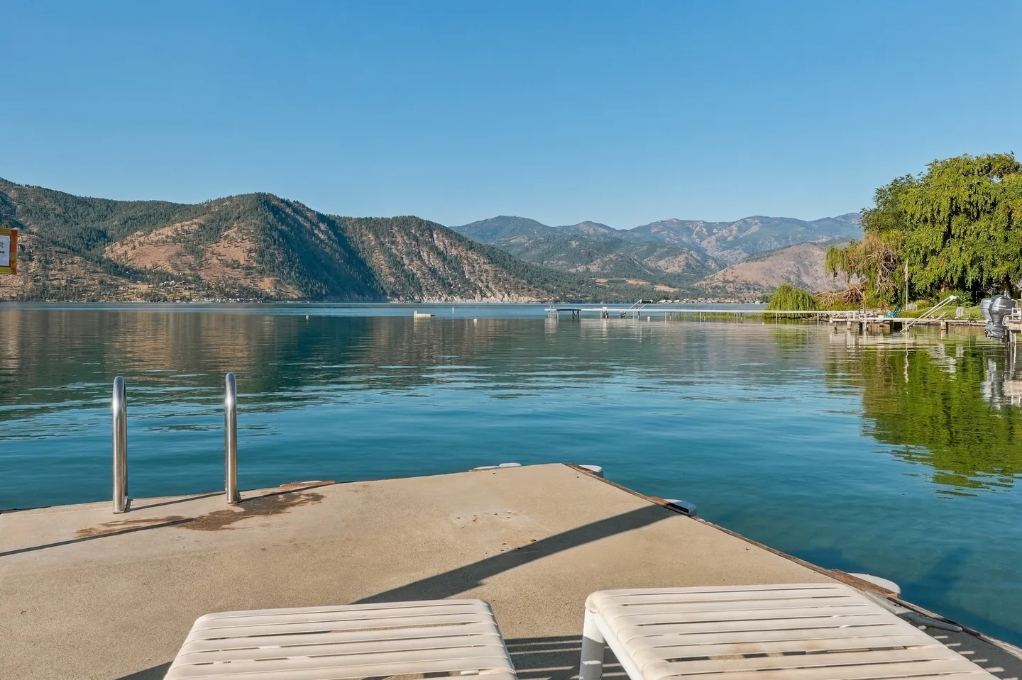 Concrete dock with a ladder and lounge chairs overlooking a calm lake surrounded by mountains under a clear blue sky.