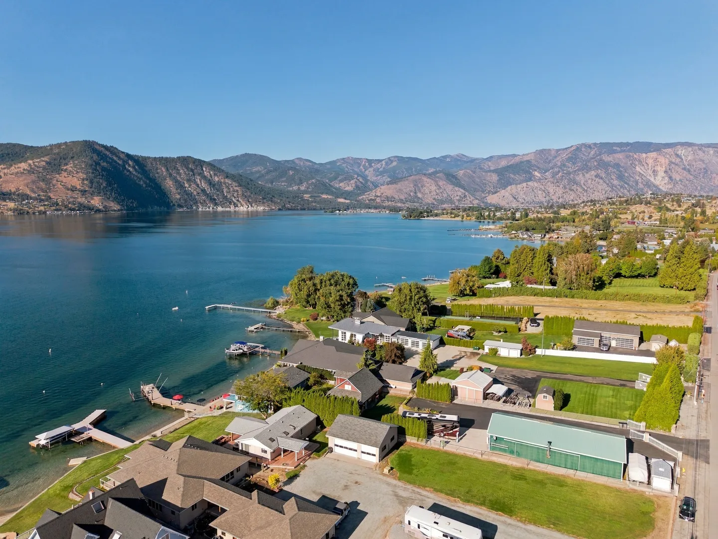 Aerial view of a lakefront neighborhood featuring residential houses, private docks, green lawns, and mountains across the water under a clear blue sky.