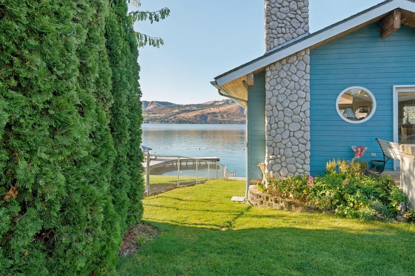 A green lawn beside a blue house with a stone chimney, leading towards a lake and dock bordered by a tall evergreen hedge.