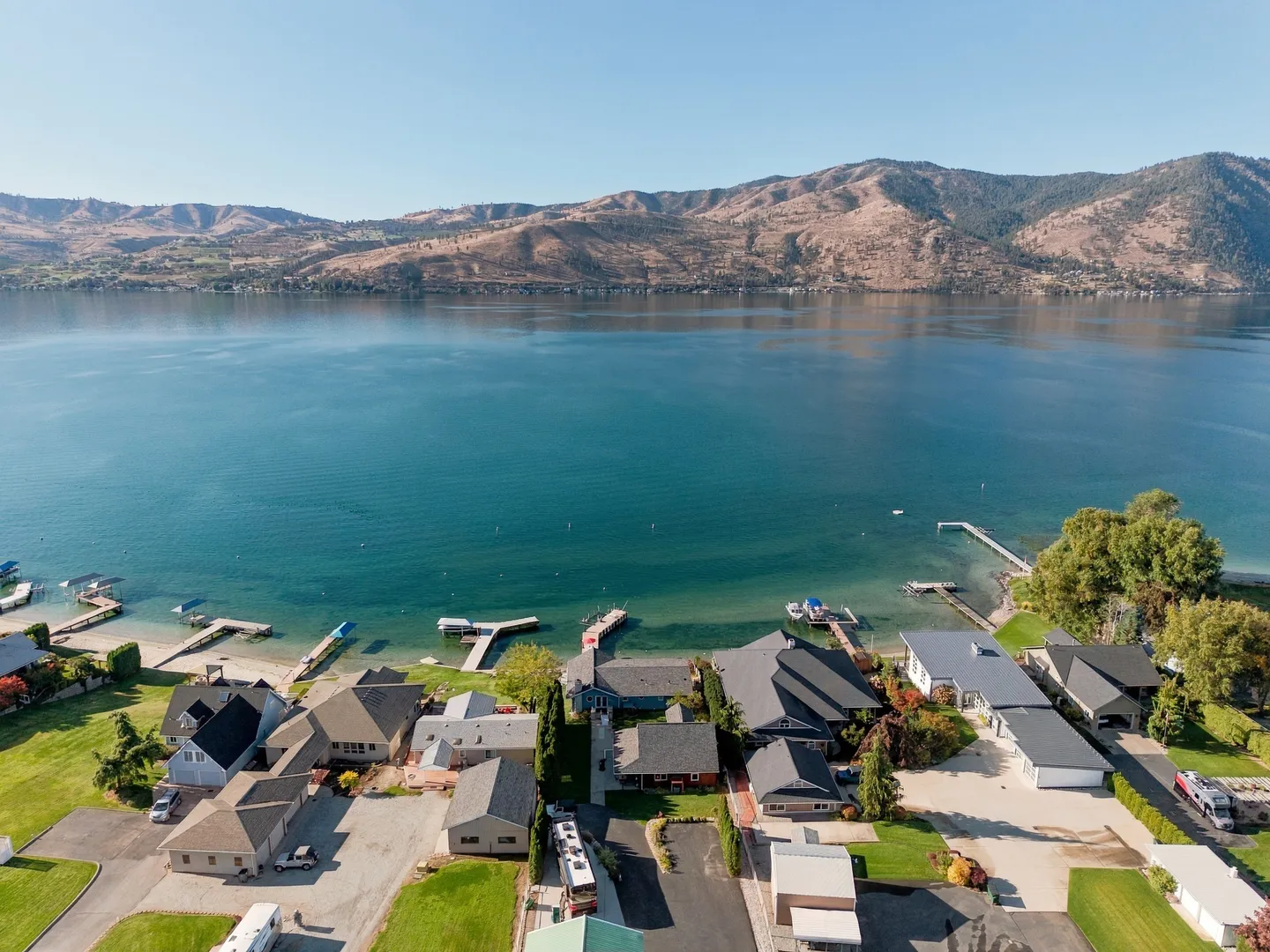 Aerial view of a lakefront residential neighborhood with docks extending into the clear blue water against a mountainous backdrop.
