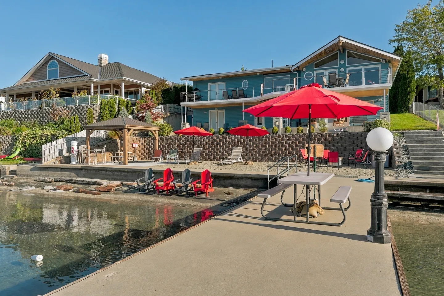 Waterfront view of a teal multi-story house with a dock, outdoor seating, red umbrellas, and a gazebo.