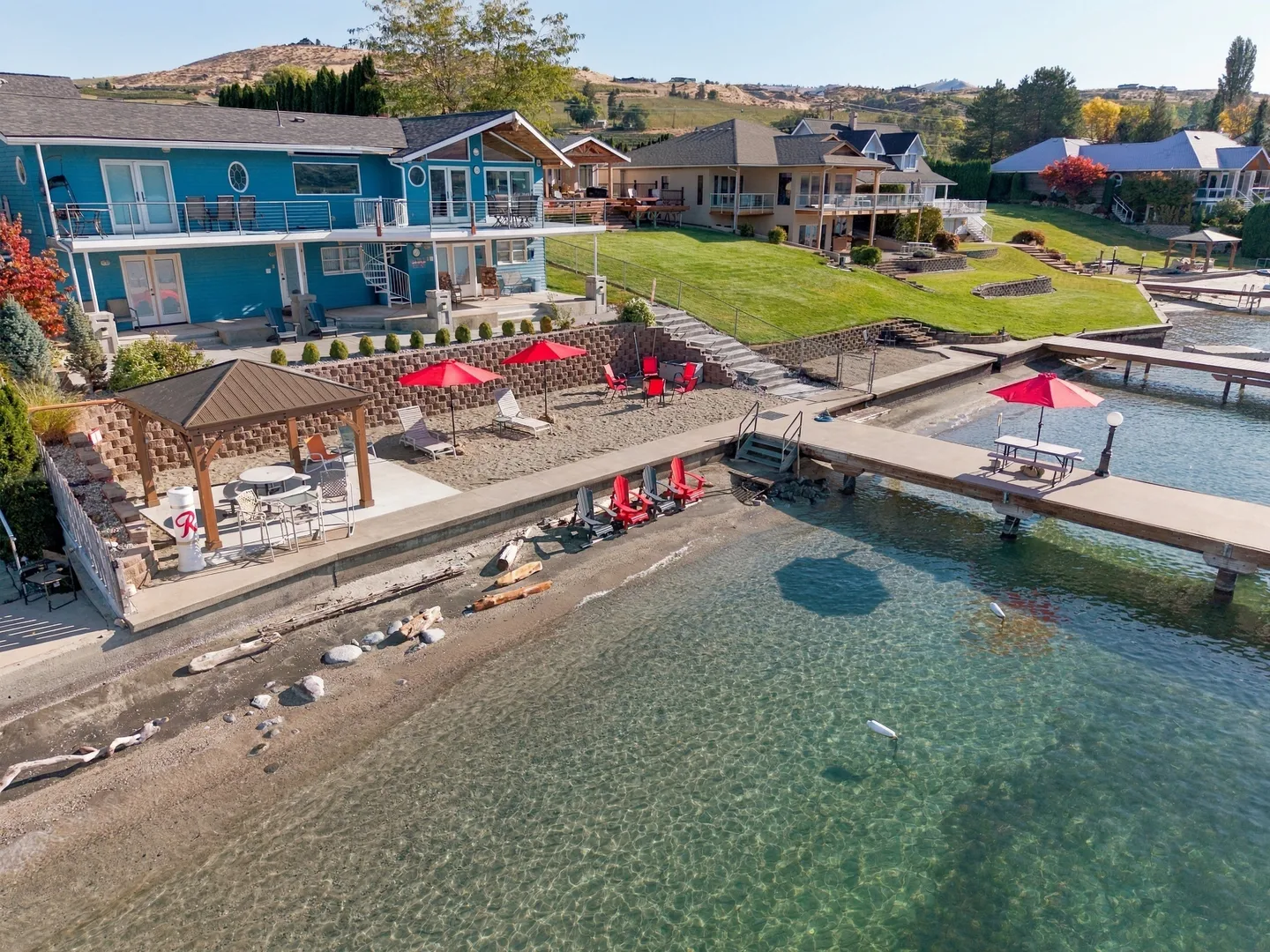 Aerial view of a blue waterfront vacation home with a terraced yard, private beach, gazebo, and dock leading into clear water.