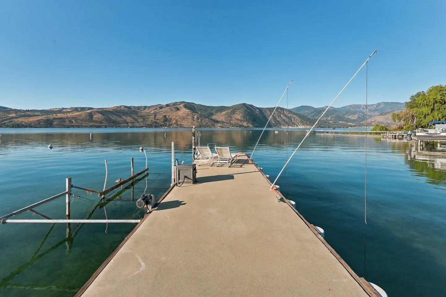 A long private dock extends into clear blue lake water, featuring lounge chairs and a view of mountains under a clear sky.