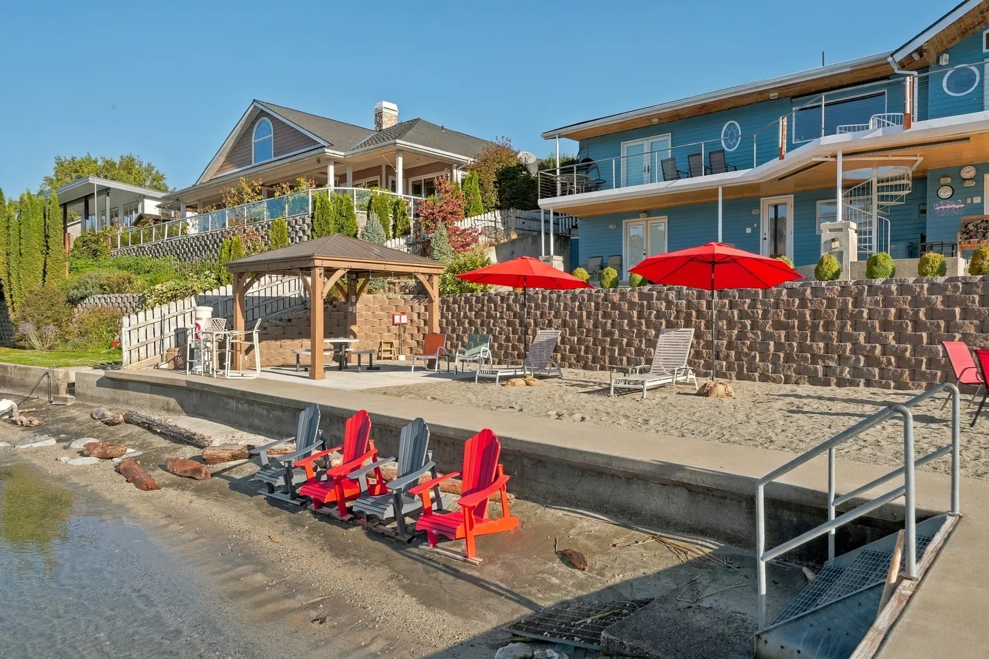 Lakeside patio and beach area with Adirondack chairs, umbrellas, gazebo, and retaining wall below houses under a clear blue sky.