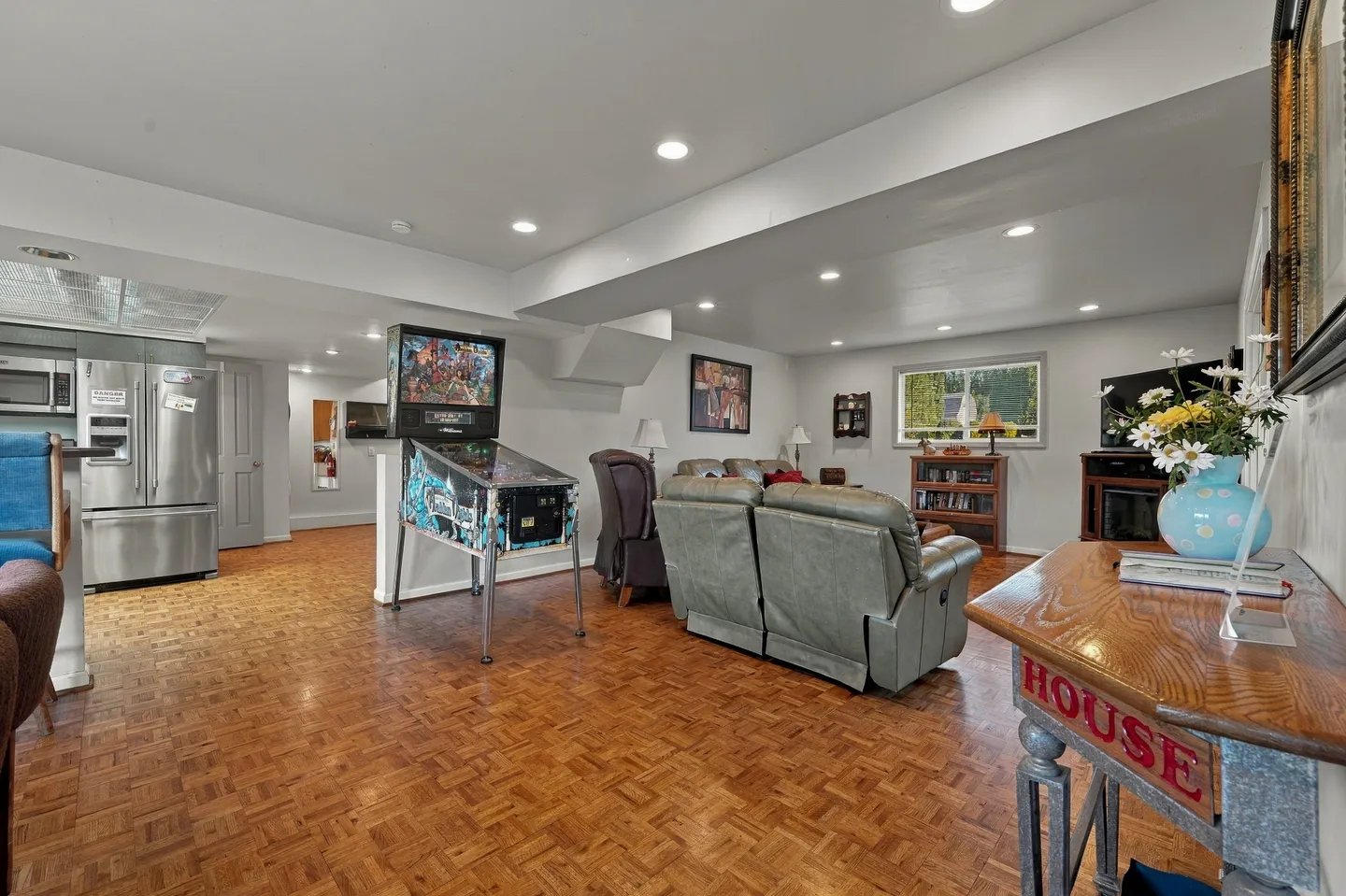 Open-concept finished basement with parquet-style flooring, a pinball machine, leather reclining sofas, and a kitchenette area.