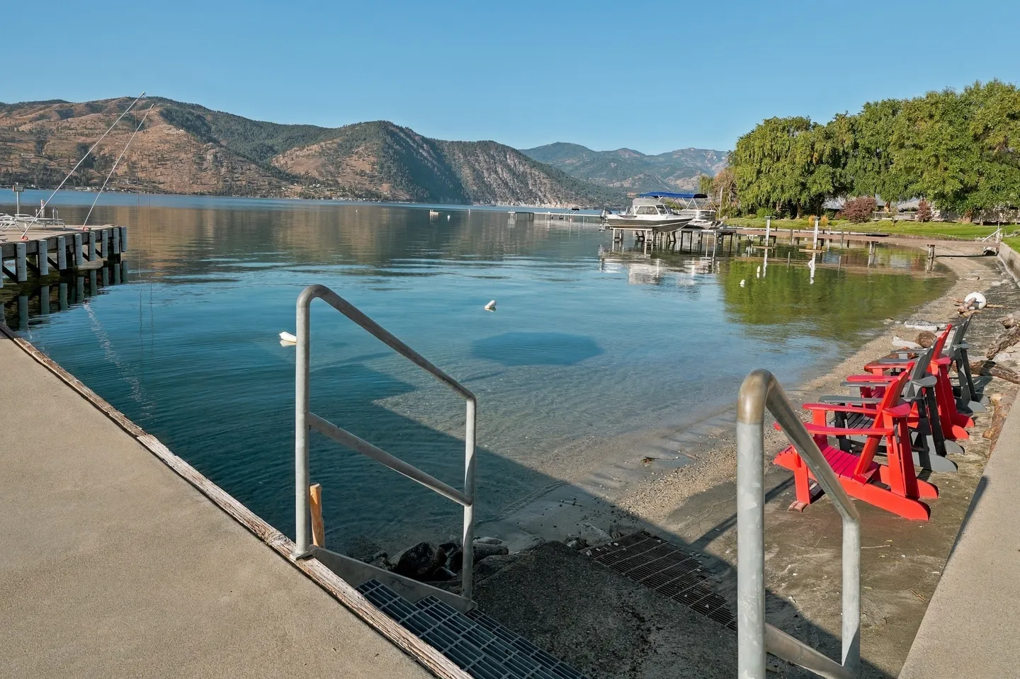 A concrete dock with handrails leading into calm, clear lake water adjacent to a small shoreline with red chairs and distant mountains.
