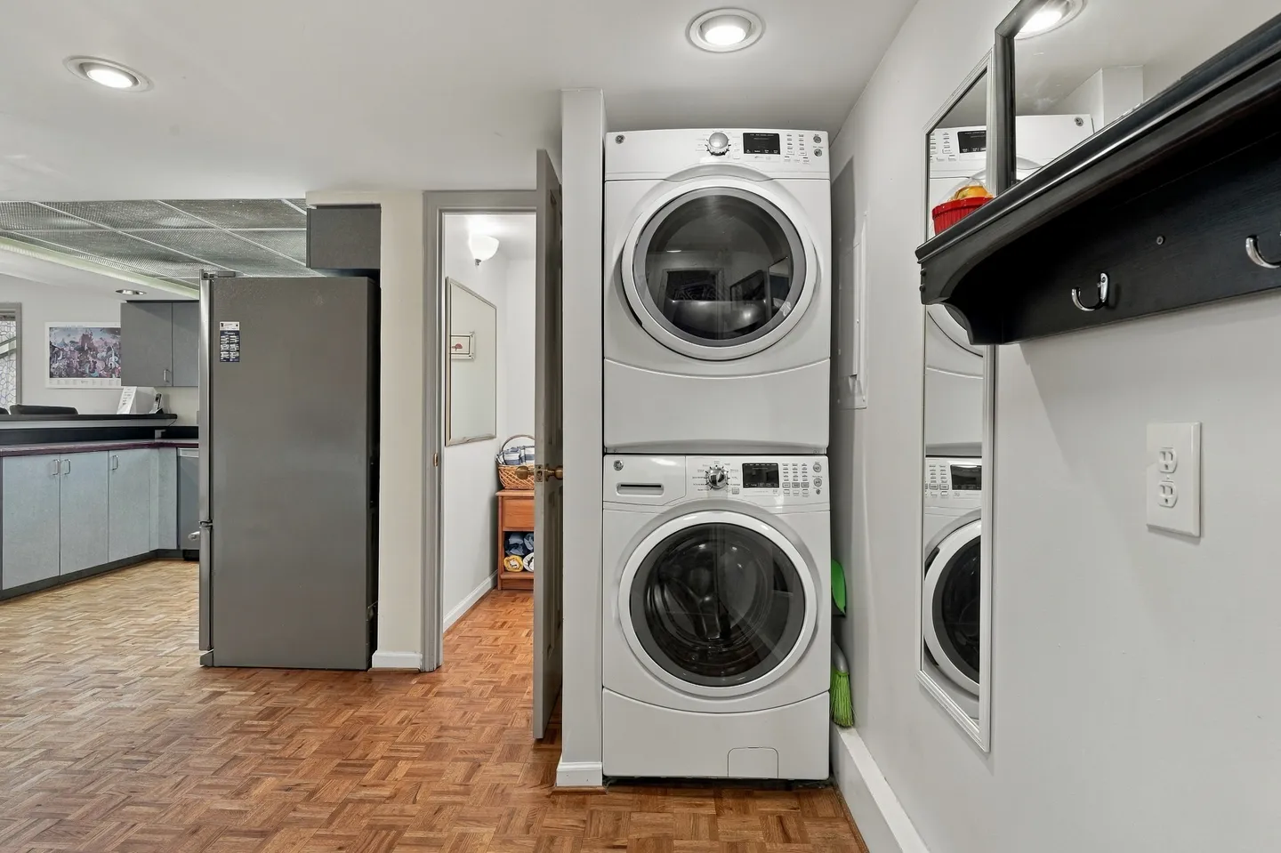 Stacked front-loading washer and dryer in a white alcove next to a parquet floor hallway and kitchen area.