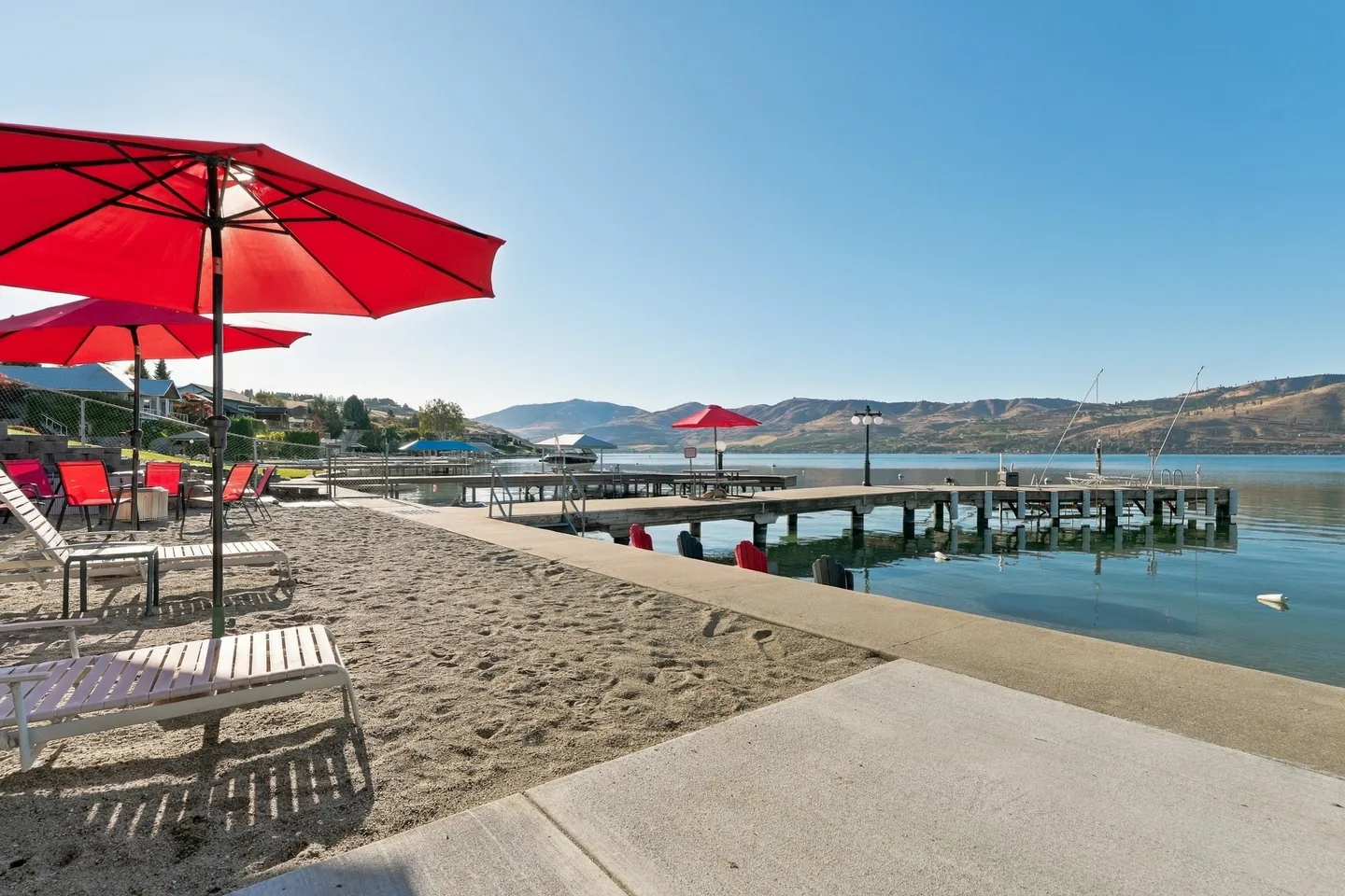 Sandy lakefront area with lounge chairs and red umbrellas next to a wooden dock and calm water with rolling hills in the background.