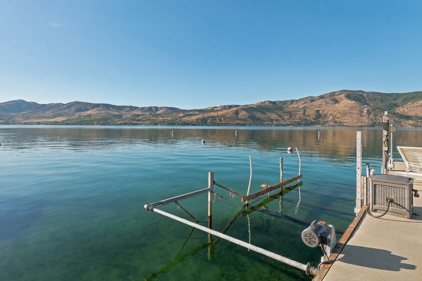 A boat lift mechanism submerged along a dock on calm, clear lake water with hills in the background under a blue sky.