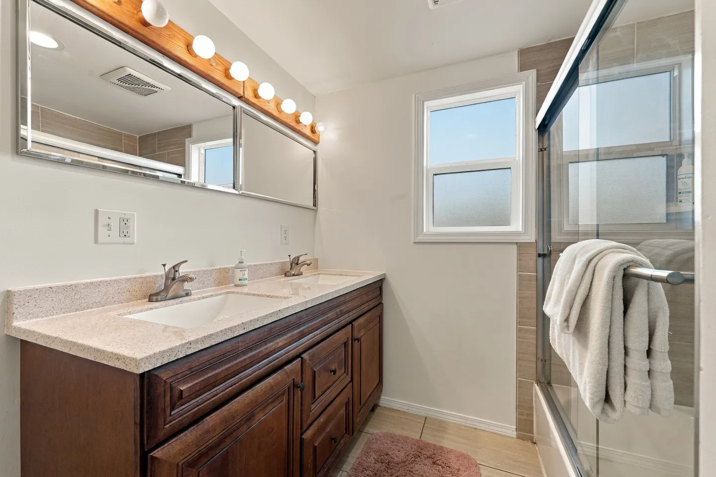 Bathroom with a double-sink dark wood vanity, illuminated mirror, and glass shower-tub enclosure.