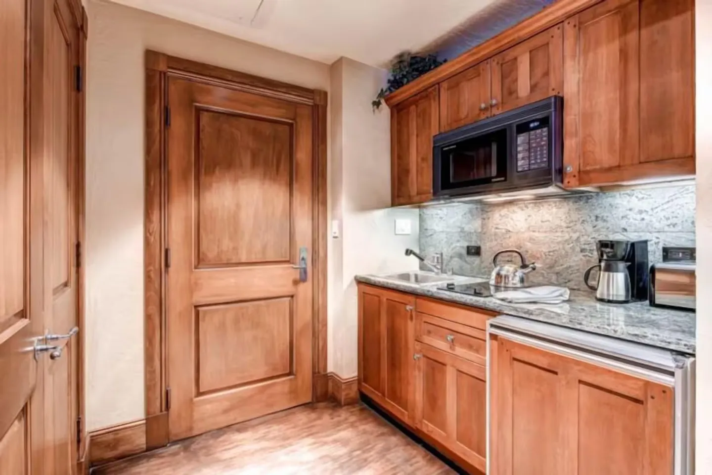 Kitchenette with wooden cabinetry, granite countertop, built-in microwave, small sink, and compact appliances next to a wooden door.