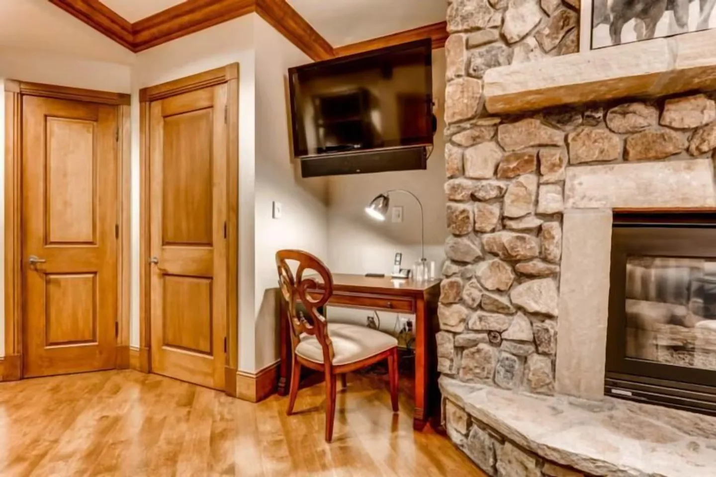 A small desk with a chair and lamp tucked into a nook beside a stone fireplace and below a wall-mounted television.