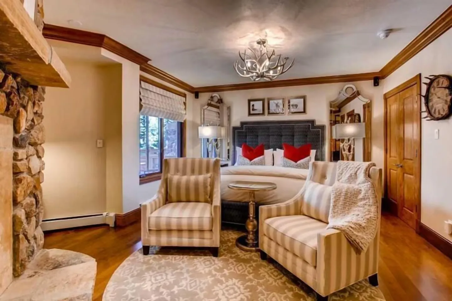 Bedroom featuring a stone fireplace, wood floors, a tufted headboard bed, and two striped armchairs around a small table.