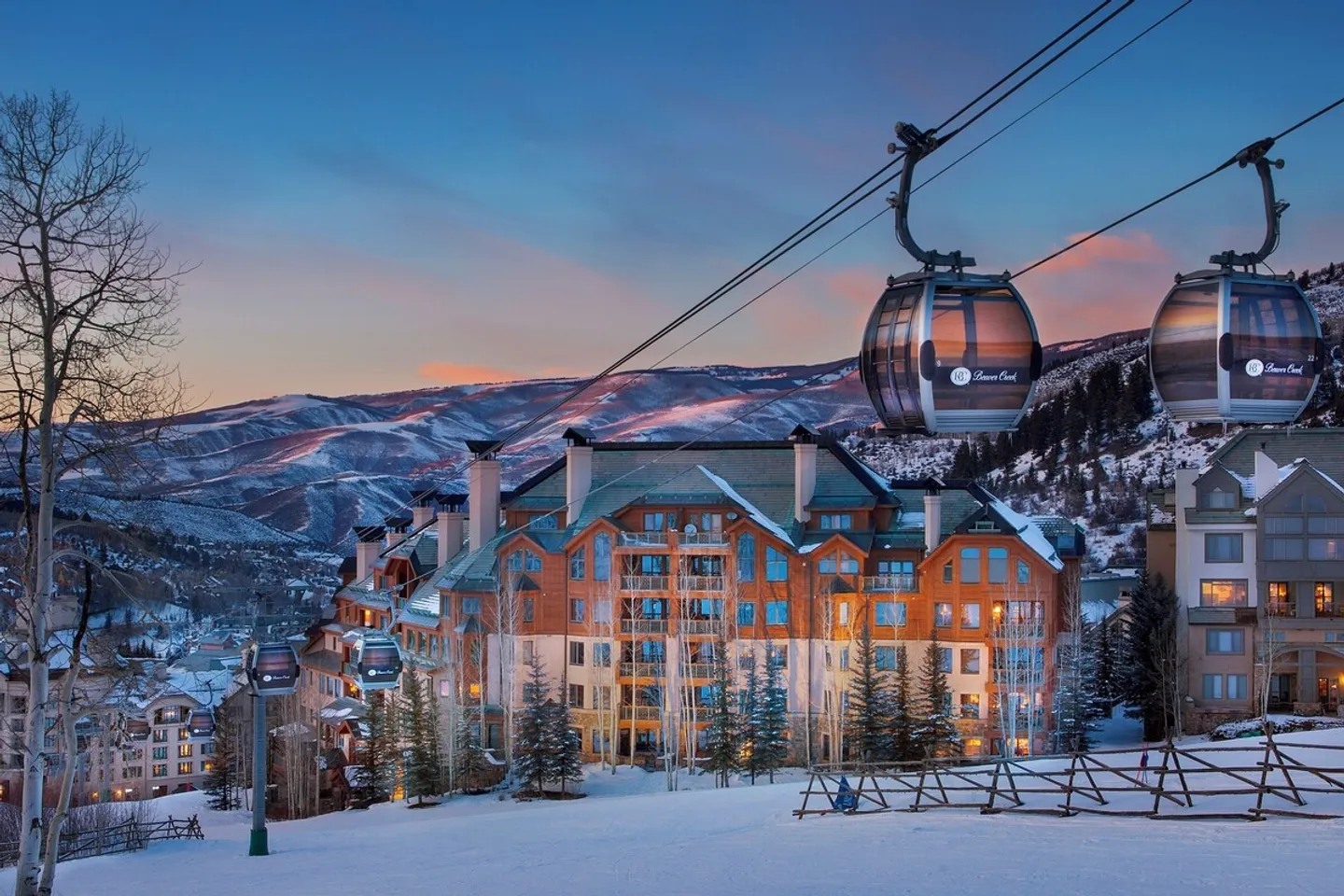 Exterior view of a large mountain ski resort lodge at dusk with gondola cabins passing overhead above snow-covered slopes.