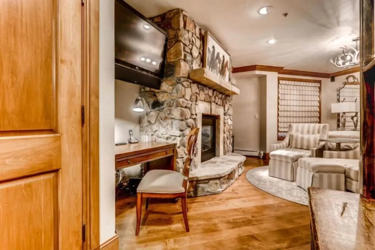 A cozy bedroom sitting area featuring a stone fireplace, a desk with a chair, and striped armchairs on wood flooring.