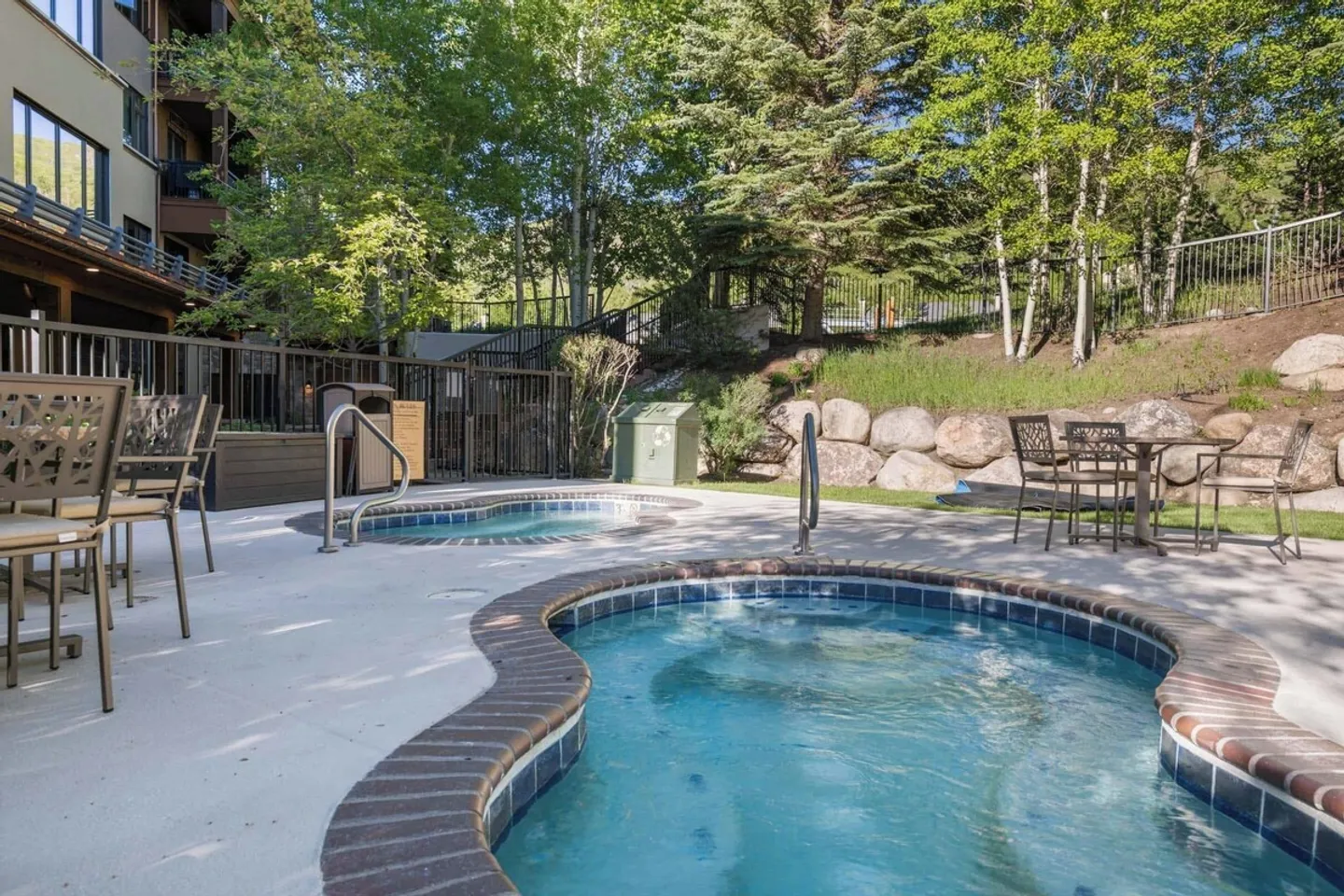 Outdoor hot tubs on a concrete patio surrounded by patio tables, chairs, metal fencing, and green trees.