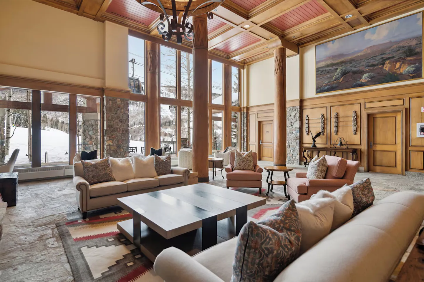 Spacious ski lodge lounge with stone tile floors, high timbered ceilings, and large floor-to-ceiling windows looking out onto snowy slopes and a ski gondola.