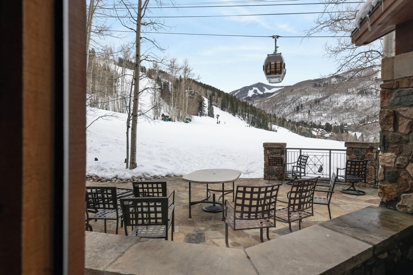 A stone outdoor patio with a table and chairs overlooking a snowy ski slope with a passing gondola.