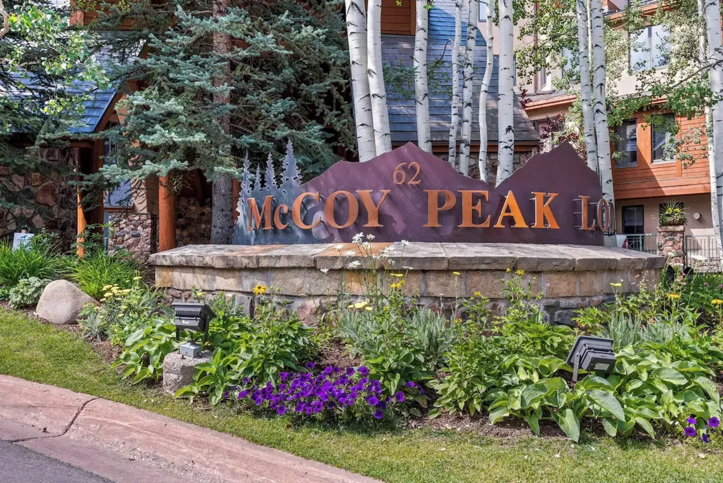 Stone entrance sign reading 62 McCoy Peak Lodge surrounded by landscaping and birch trees.