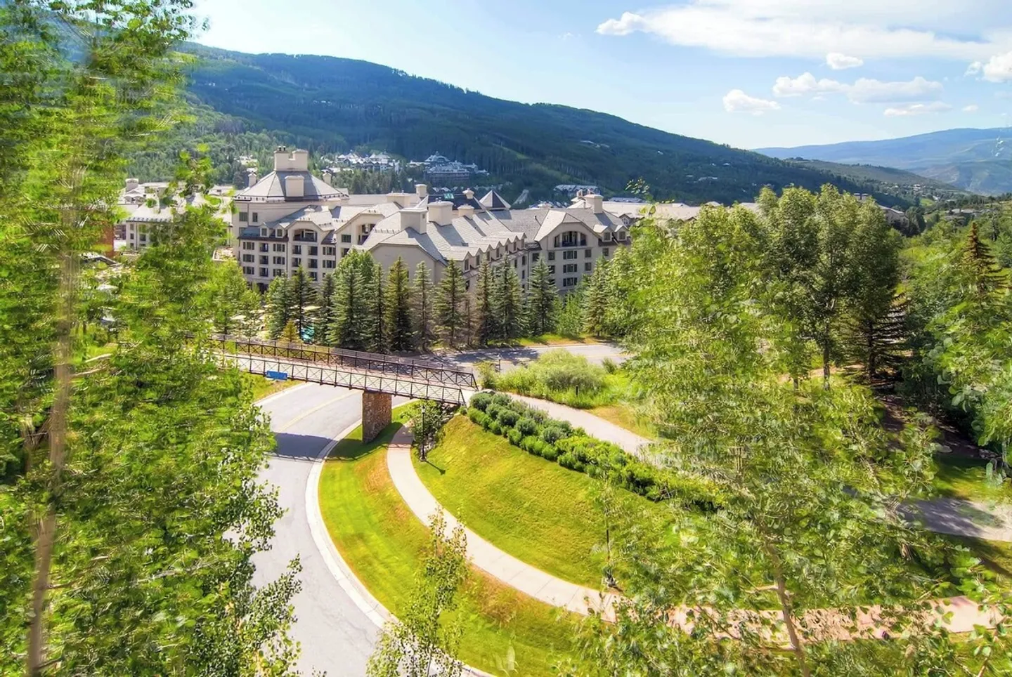 Elevated view through birch trees overlooking a paved road, pedestrian bridge, and large alpine resort building backed by forested mountains.