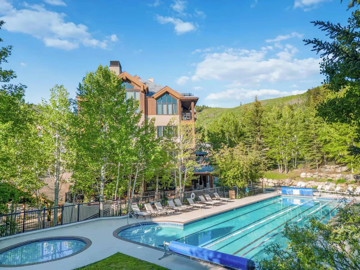 Outdoor swimming pool with lap lanes and round hot tub next to a multi-story lodge surrounded by trees and a hillside.
