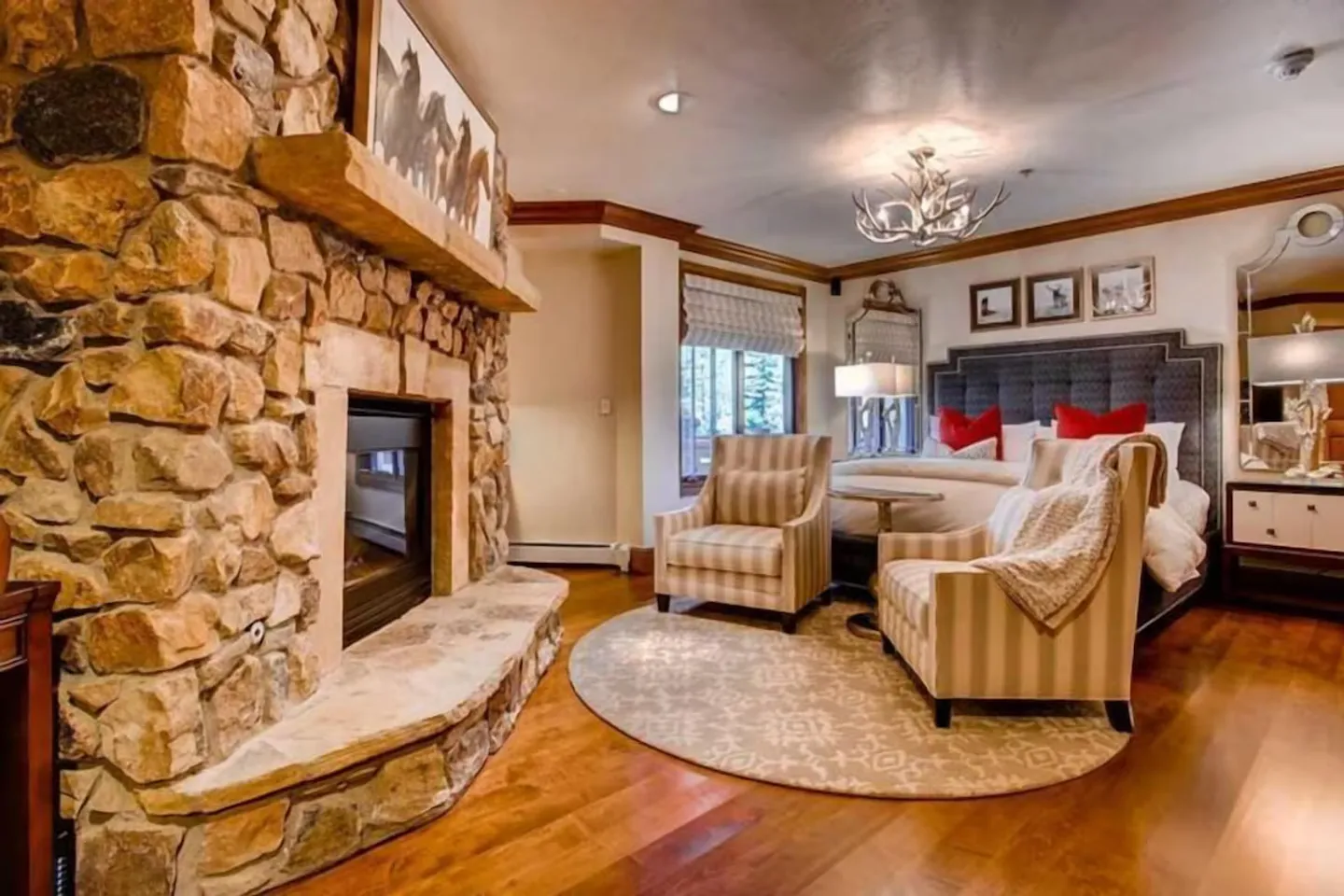 Bedroom featuring a large stone fireplace, hardwood floors, a bed with a tufted headboard, and two armchairs on a round area rug.