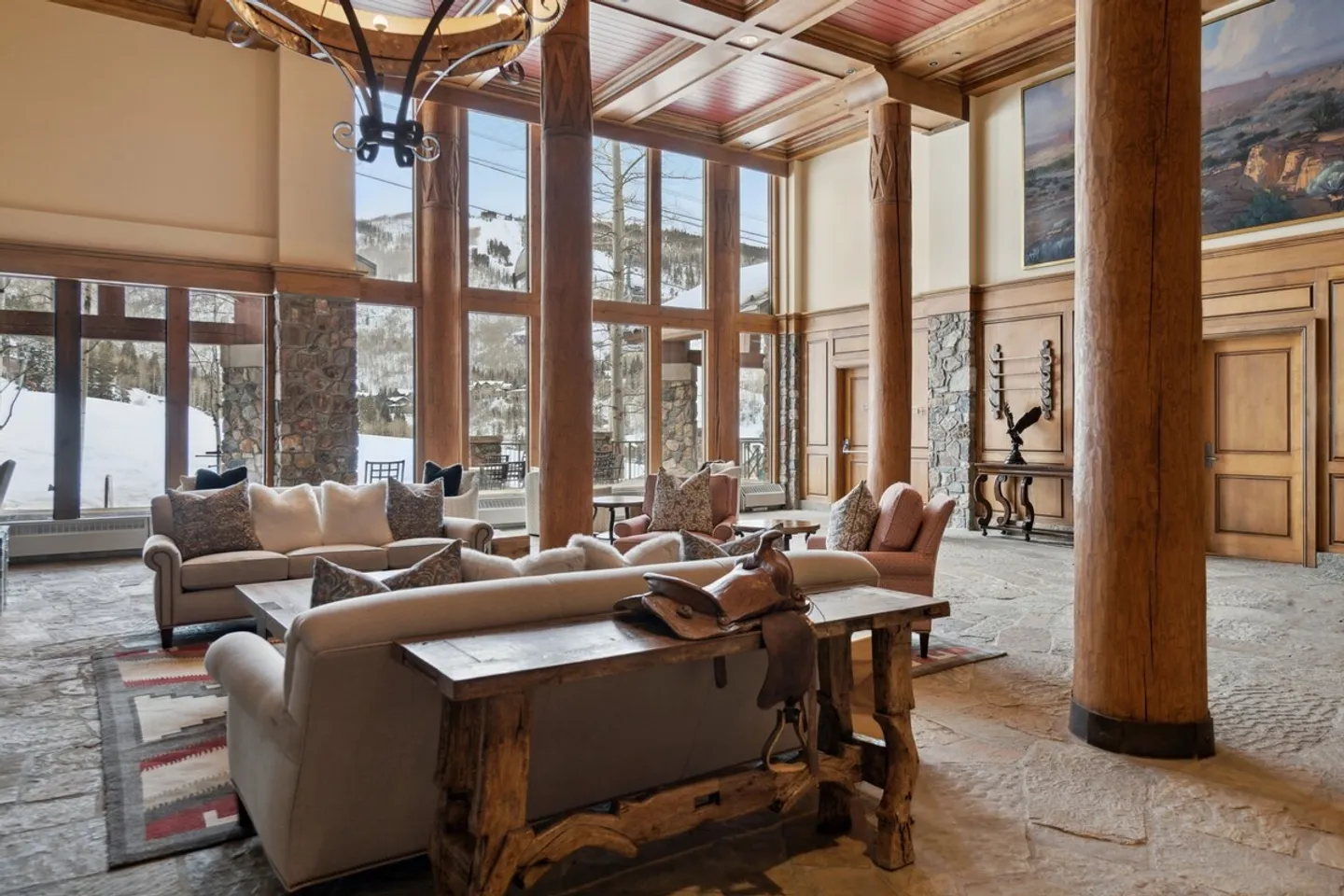 A spacious lodge living room with massive wood log columns, stone flooring, couches, and floor-to-ceiling windows showing a snowy mountain view.