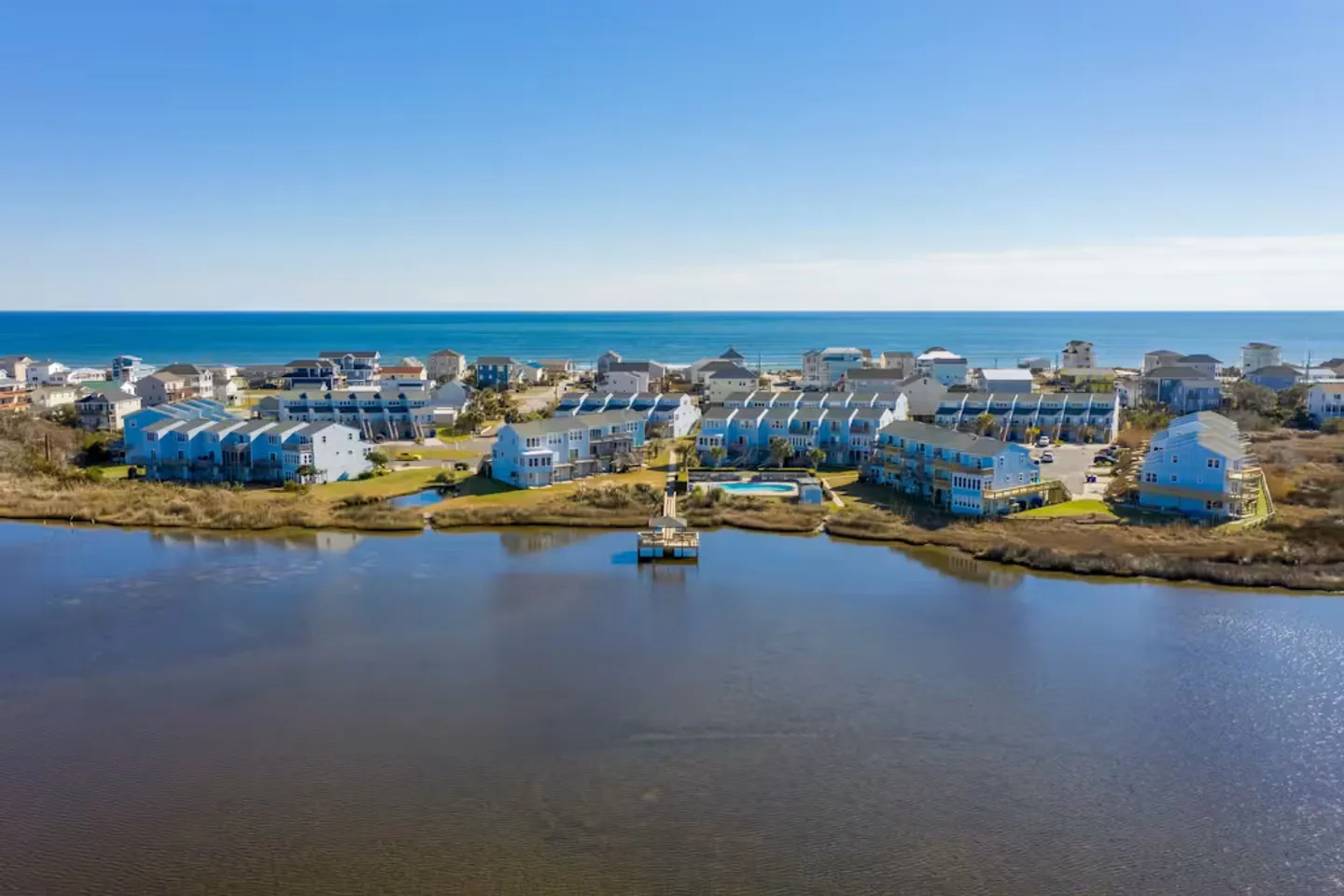 Aerial view of a coastal neighborhood with blue multi-story residential buildings bordered by an inlet and the ocean in the background under a clear sky.
