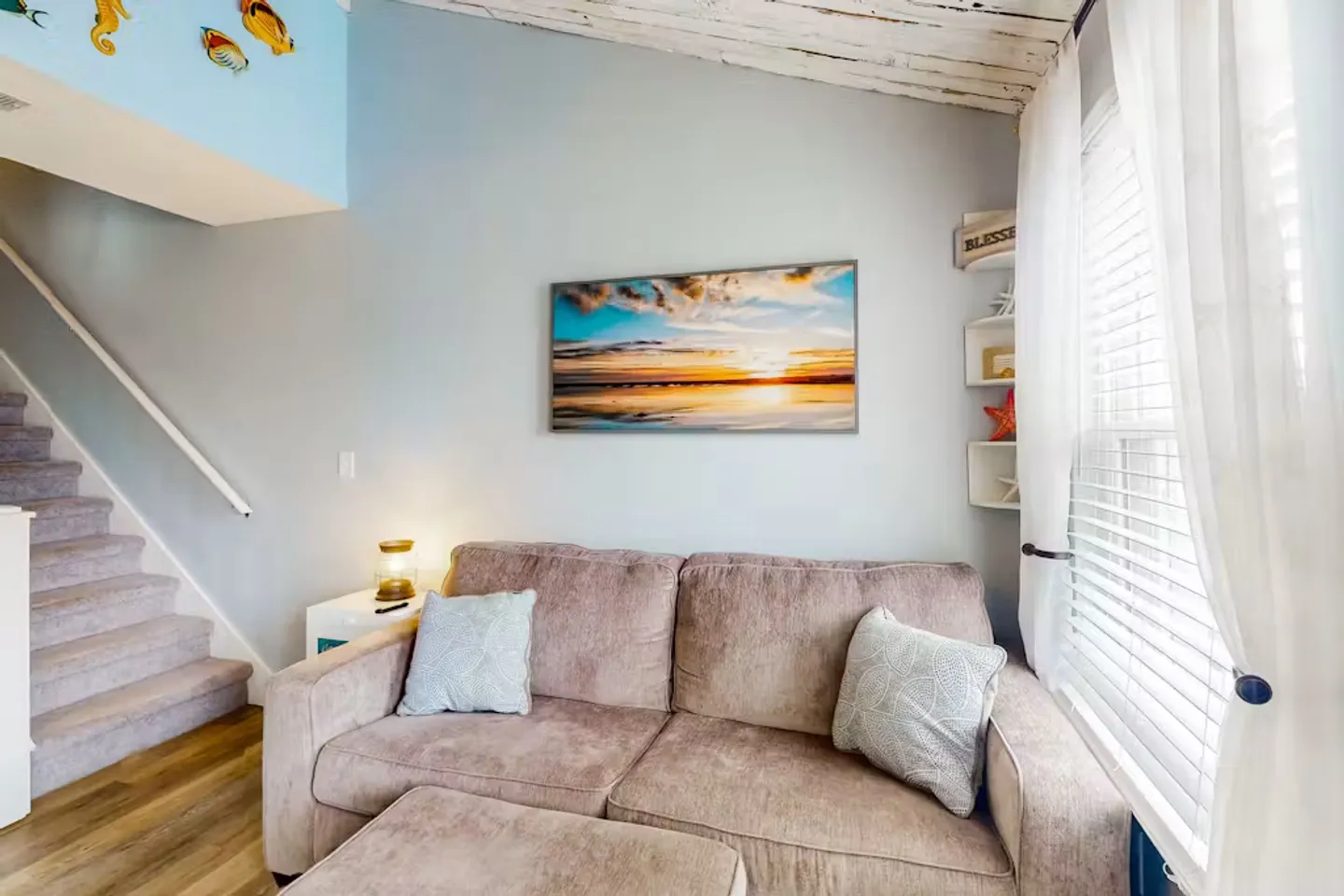 Living area featuring a plush sofa, matching ottoman, coastal sunset artwork, and carpeted stairs leading to the upper level.
