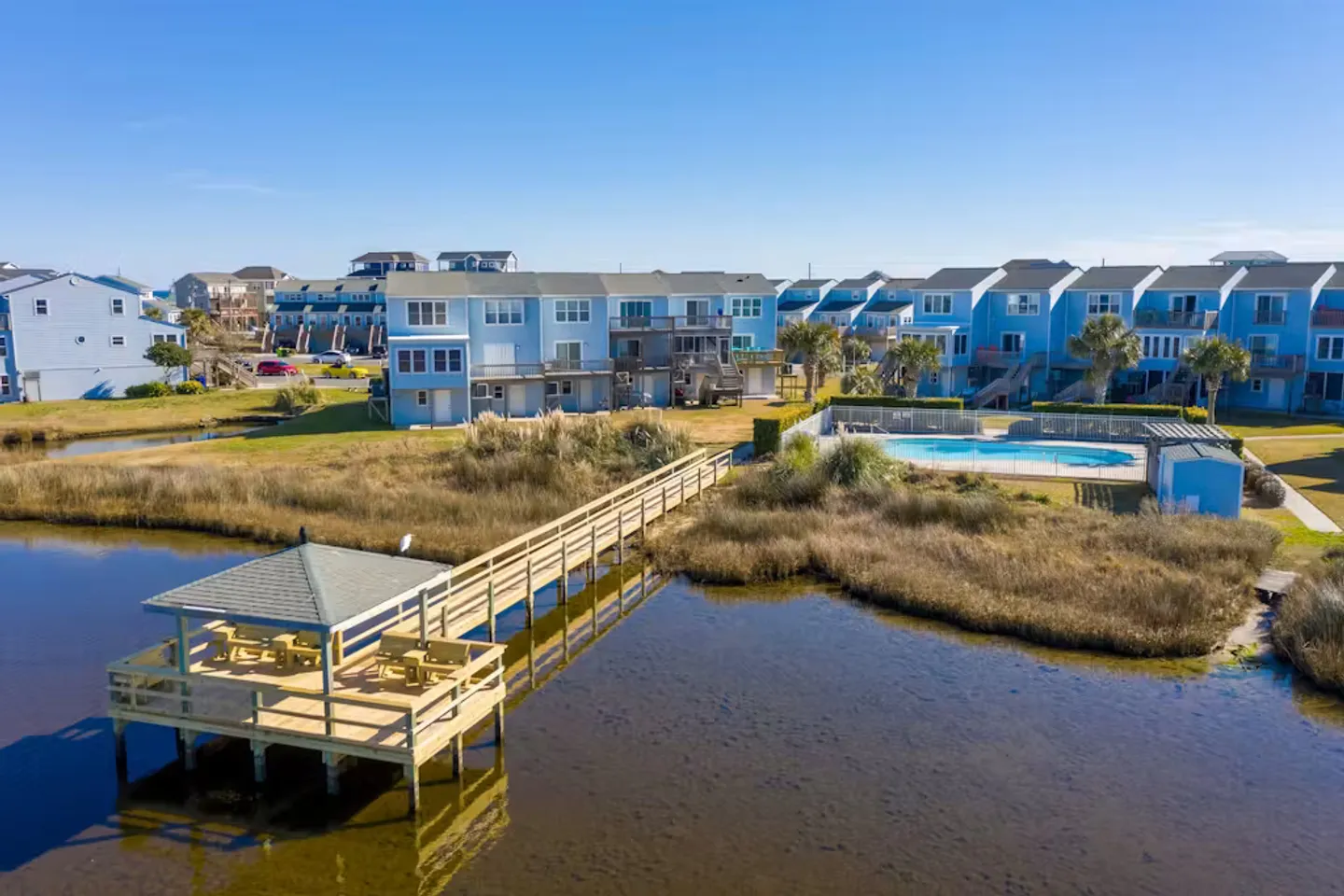 Aerial view of a coastal townhouse complex with a wooden pier and gazebo over the water, alongside a fenced outdoor pool.