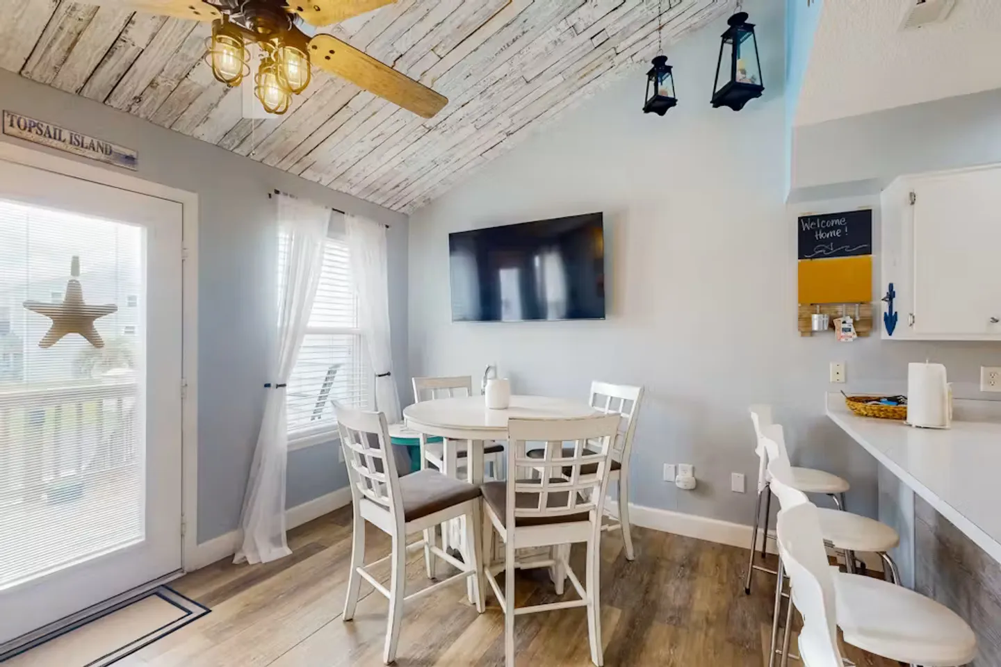 Dining area with a round white table, four chairs, a wall-mounted TV, and a wood-plank style ceiling.