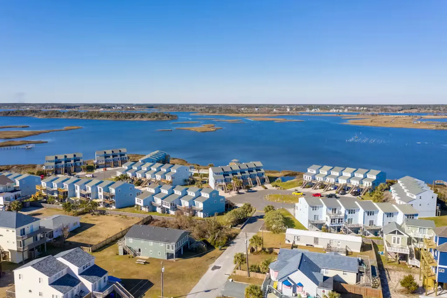 Aerial view of a coastal neighborhood with blue-sided townhomes and houses next to a large bay under a clear blue sky.