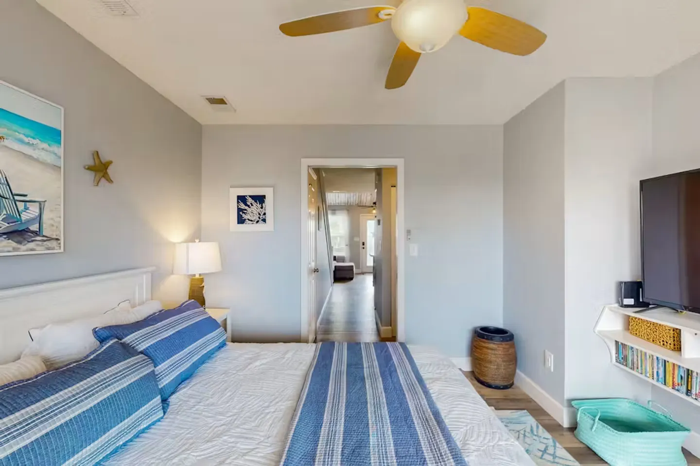 Bedroom with blue and white striped bedding, a ceiling fan, wall-mounted television, and a doorway looking down a hallway.