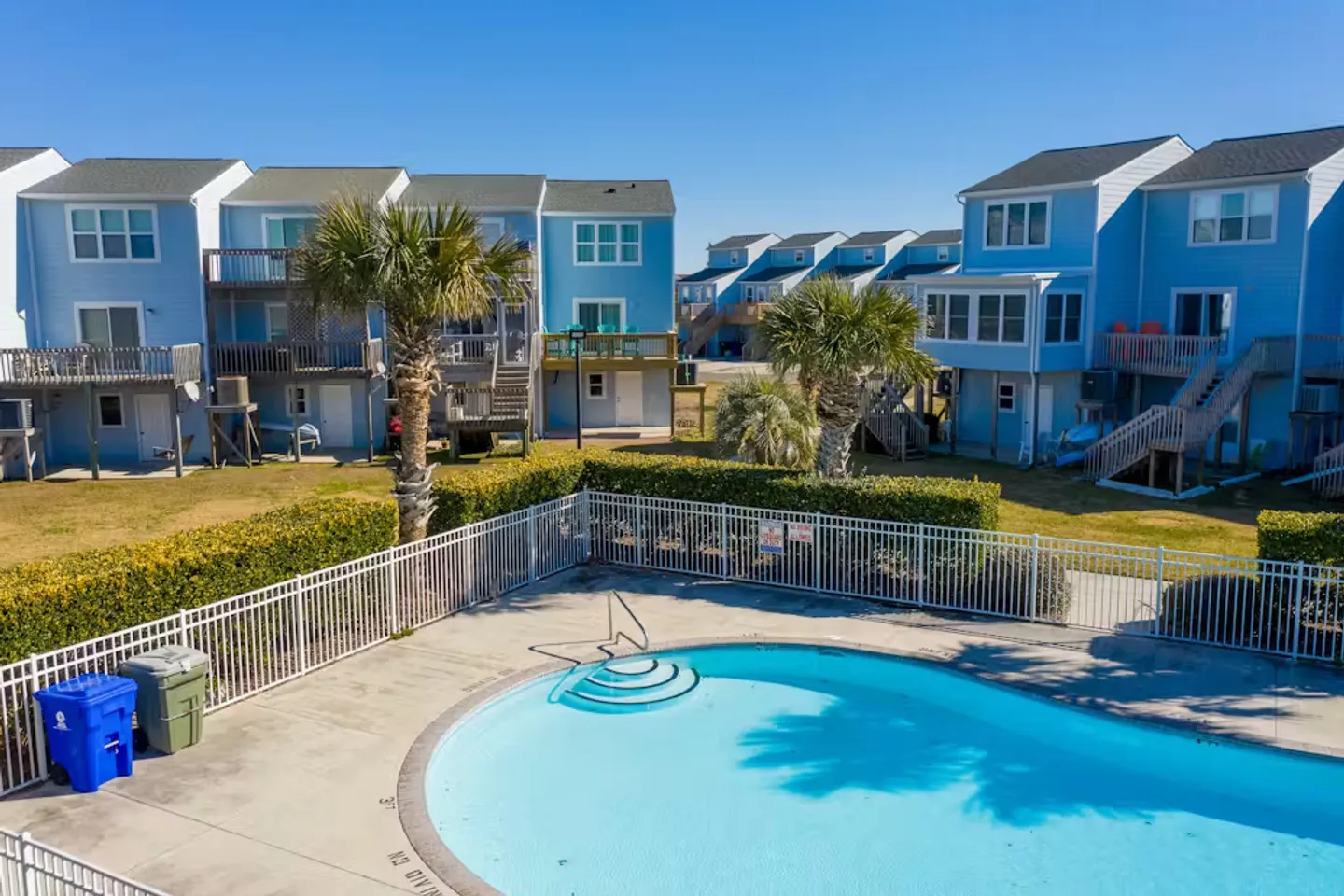 Elevated view of a fenced outdoor community swimming pool bordered by palm trees and blue multi-story beach houses.
