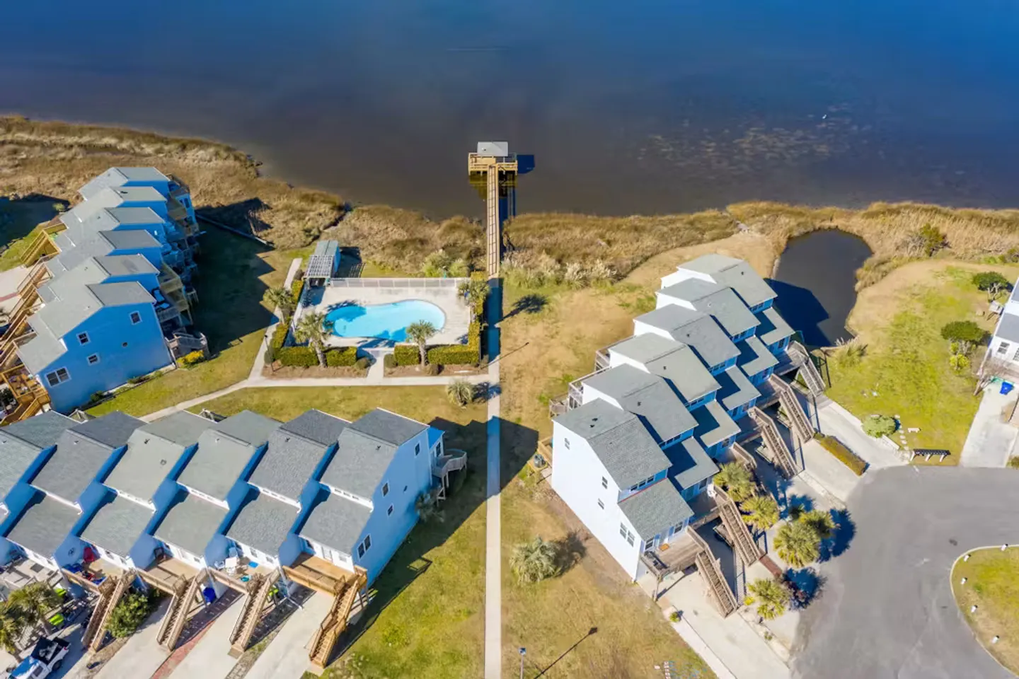 High aerial view of a waterfront townhouse community featuring rows of blue buildings, an outdoor pool, and a pier extending into the water.