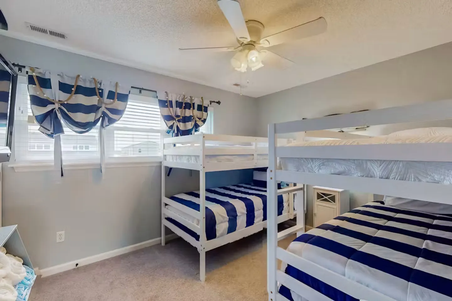 A bedroom with two white wooden bunk beds dressed in blue and white striped bedding, carpeted flooring, and a ceiling fan.