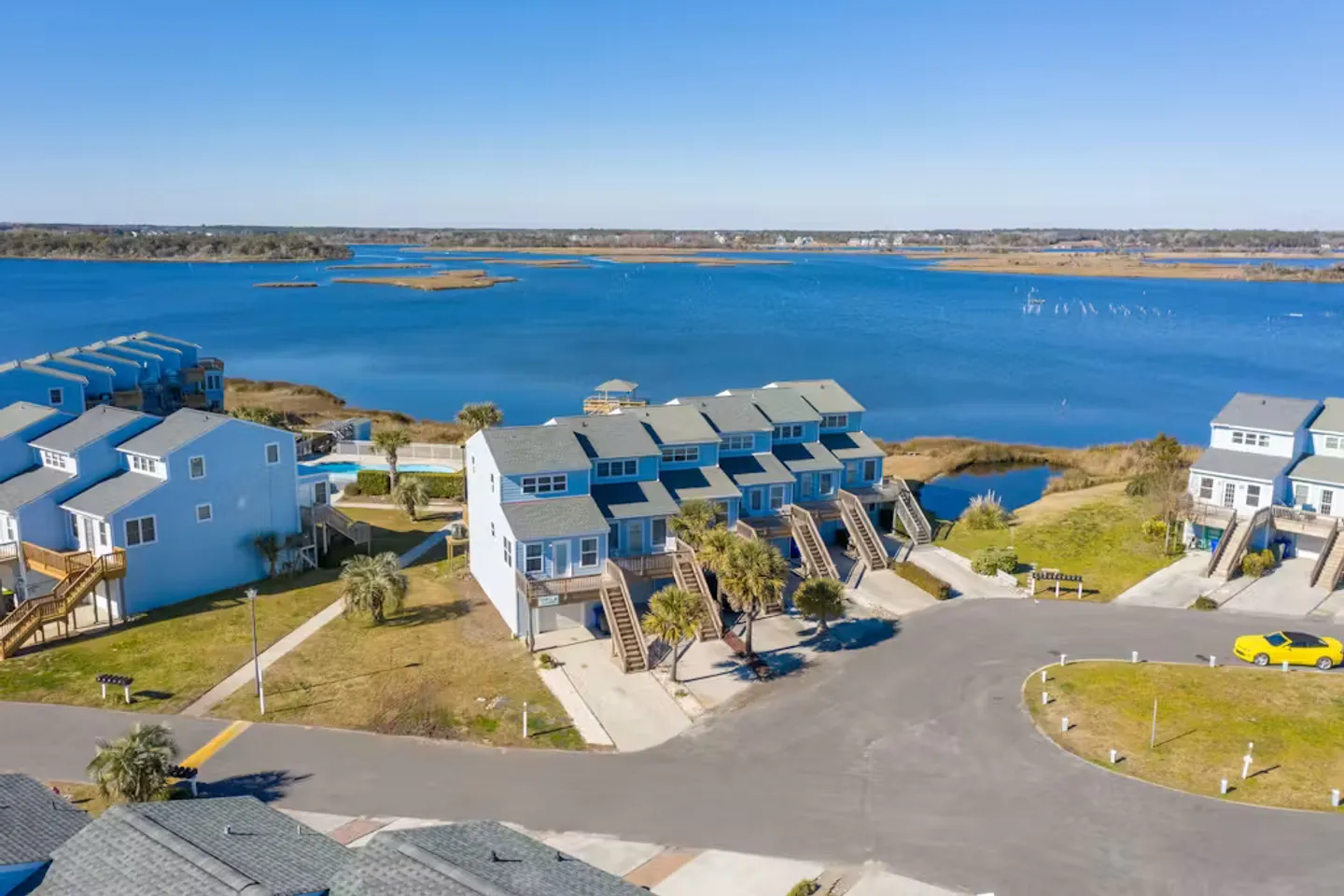 Aerial view of light blue coastal townhomes bordering a wide body of water under a clear blue sky.