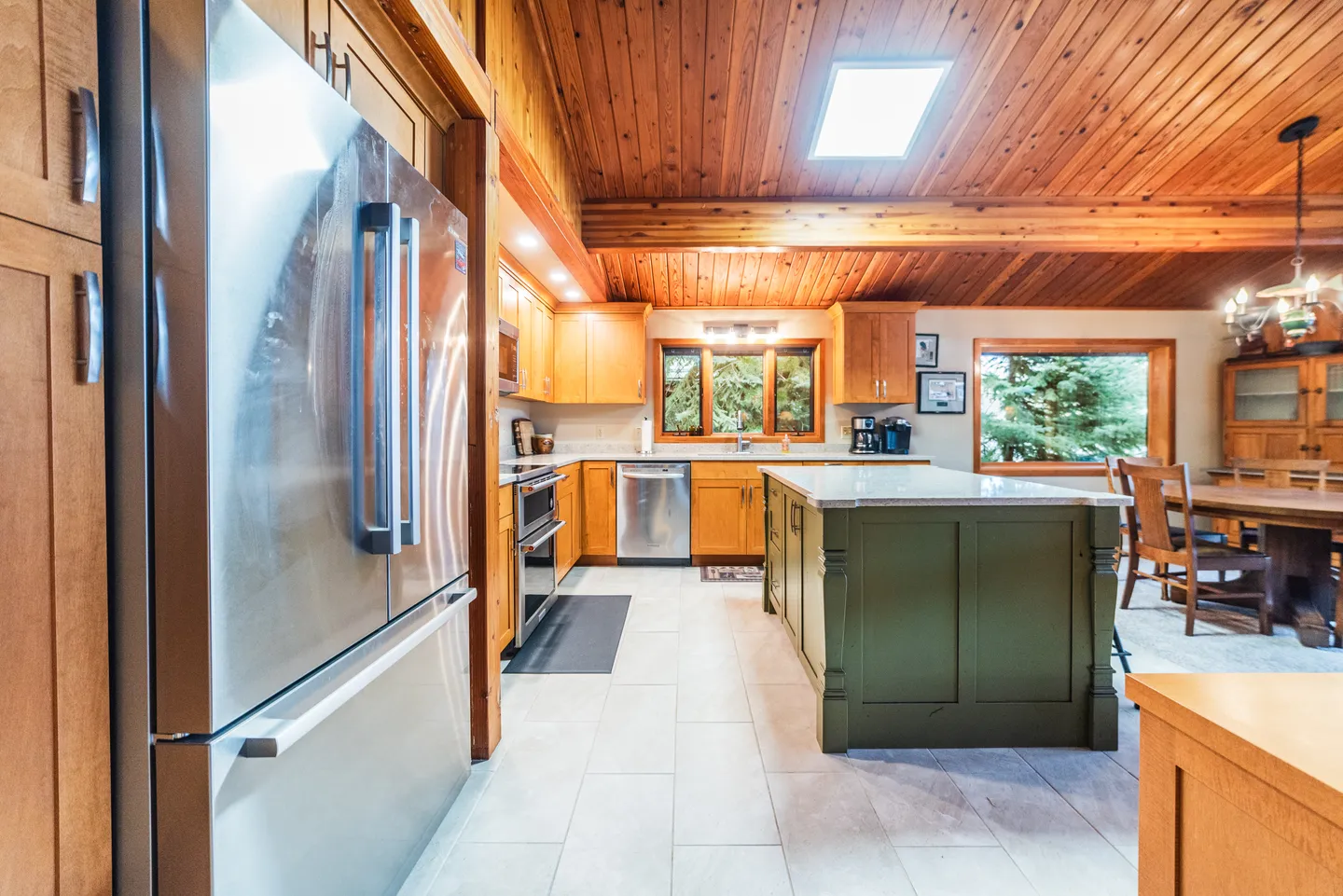 Kitchen with wood-paneled ceiling, skylight, green island, stainless steel appliances, and adjacent dining area.