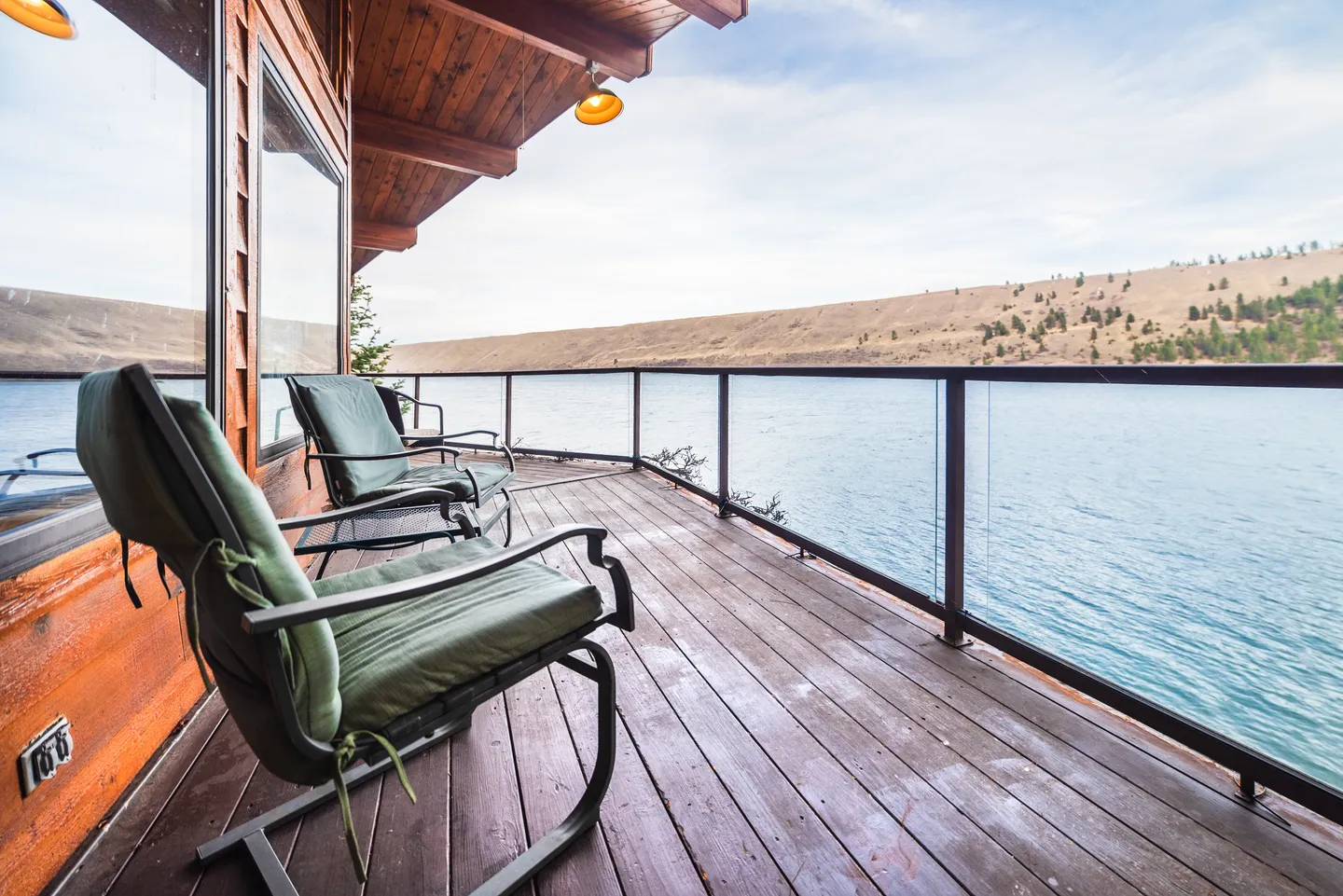 A wooden deck with two cushioned chairs and a glass railing overlooking a large lake and distant hills.