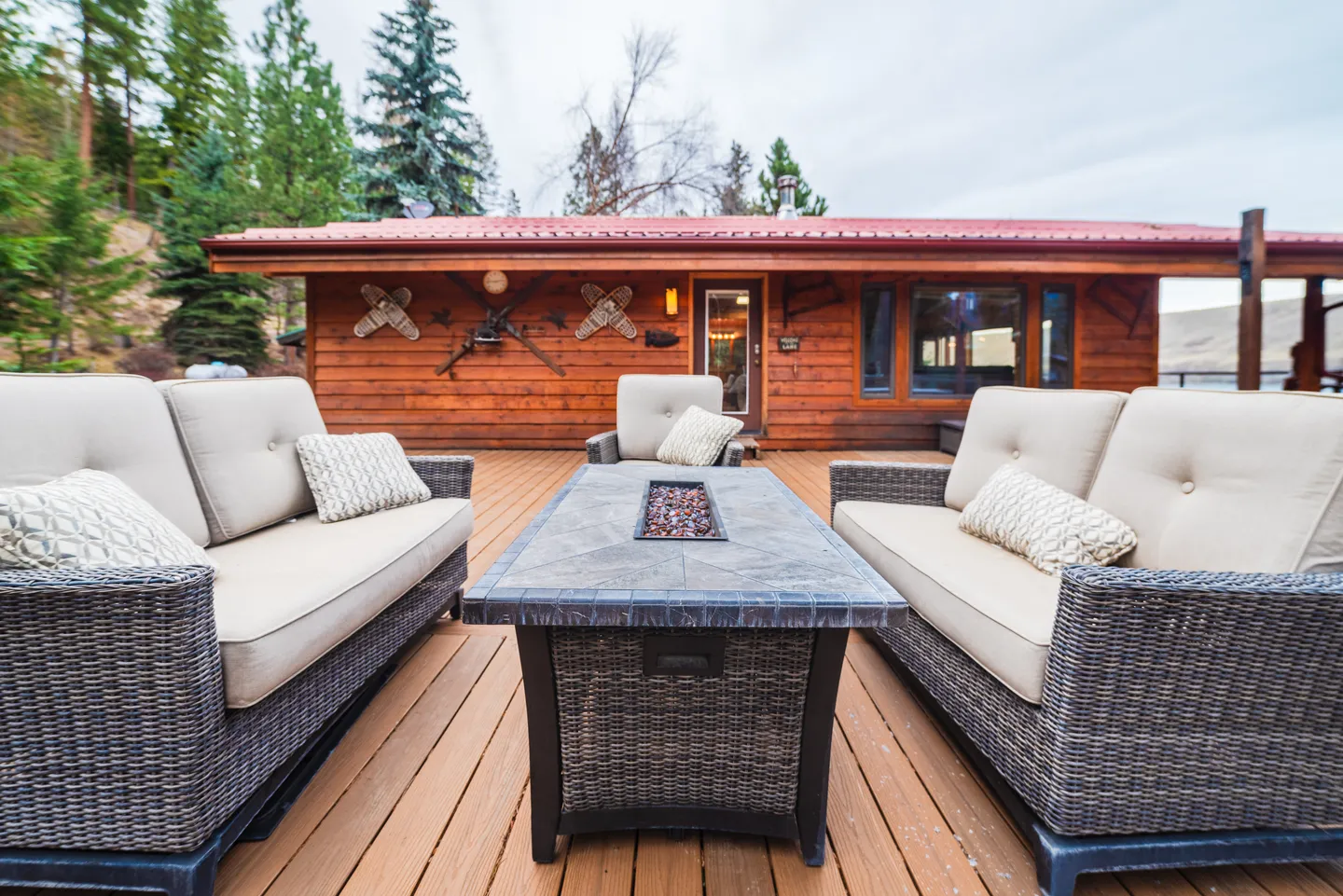 Outdoor wooden deck with wicker sofas, armchairs, and a central fire table outside a log-sided cabin.