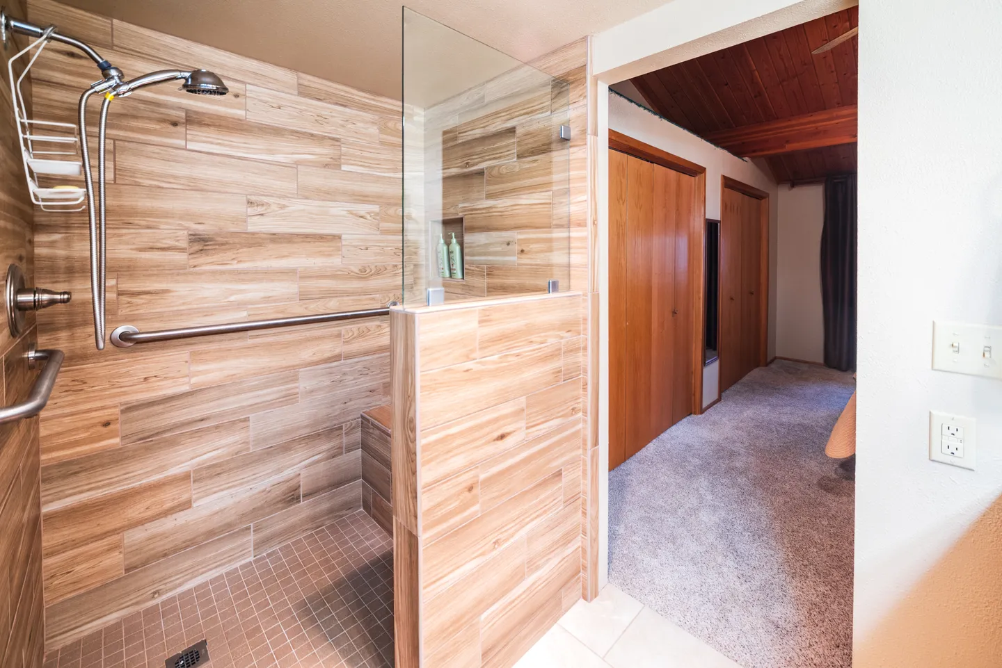 Walk-in shower with wood-look tile, glass partition, grab bars, and an adjoining carpeted bedroom area.