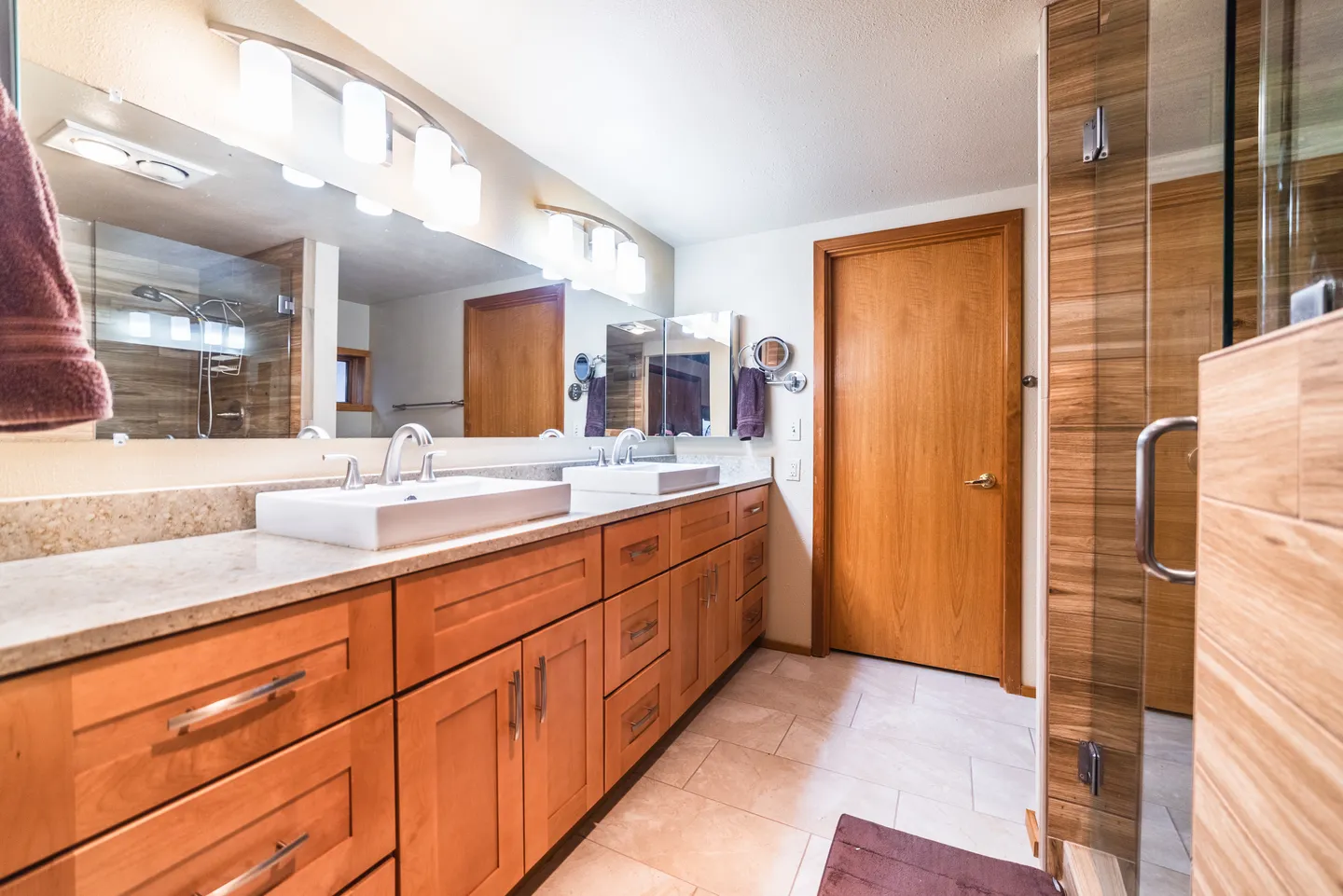 Bathroom with a long wooden double vanity, two vessel sinks, a large mirror with modern light fixtures, tiled floors, and a glass-enclosed shower on the right.