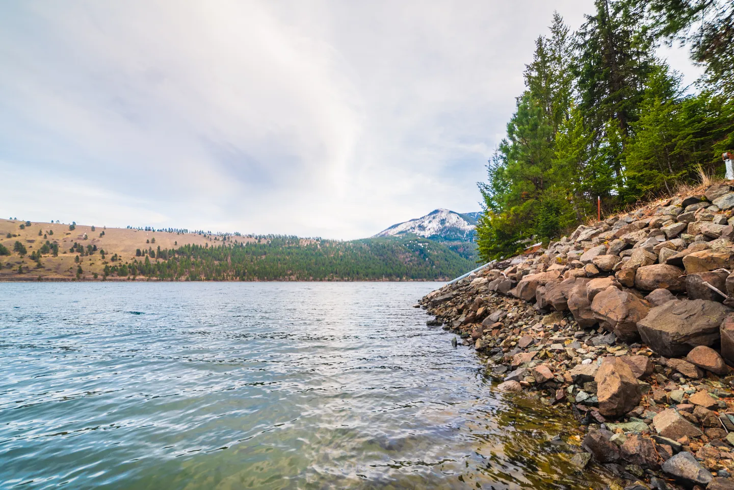 A wide view of a lake with a rocky shoreline lined with pine trees and hills in the distance under a blue sky.