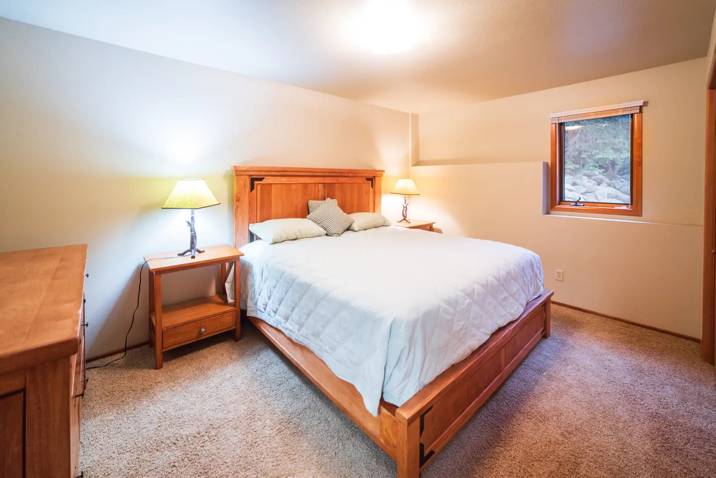 Bedroom with a wooden bed, light blue bedspread, matching wooden nightstands with lamps, carpeted floor, and a window.