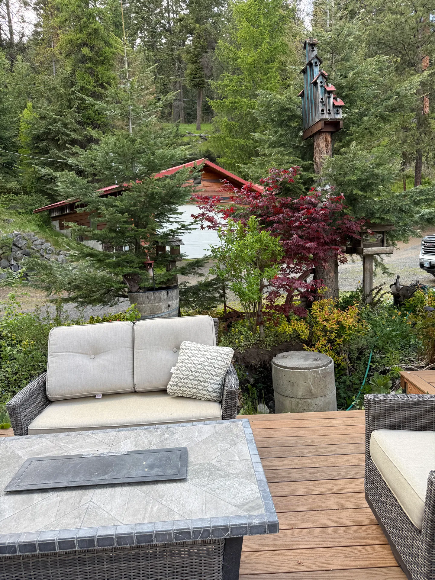 Outdoor wood deck featuring wicker seating, a tiled fire table, and views of lush trees, a red-roofed garage, and a tall decorative birdhouse.